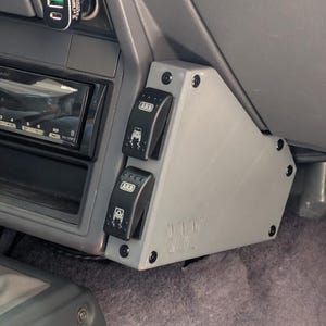 Könnte beinhalten: Eine graue Kunststoffplatte mit zwei schwarzen ARB-Schaltern, die im Innenraum eines Fahrzeugs installiert sind. Die Platte ist in der Nähe des Armaturenbretts montiert und trägt den Text "J117". Die Schalter haben beleuchtete Symbole.