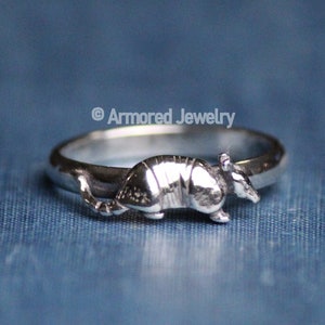 Puede incluir: Anillo de plata con un diseño de armadillo detallado. El armadillo está esculpido de forma realista, con su caparazón y cabeza visibles. El anillo está sobre un fondo azul. El texto "Armored Jewelry" es visible.