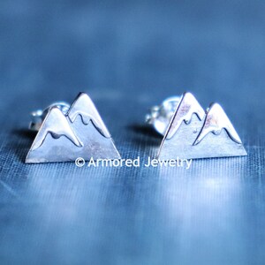 May include: A pair of silver mountain-shaped stud earrings. The earrings are small and delicate, and they feature a simple design of two mountains.