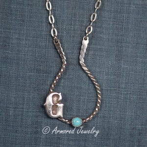 May include: Silver-toned necklace featuring a cursive letter "G" pendant and a small turquoise-colored bead. The necklace has a twisted rope design and a delicate chain. The text "Armored Jewelry" is at the bottom.