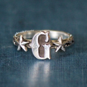May include: Silver ring with a large letter G and two star-shaped accents on the band.