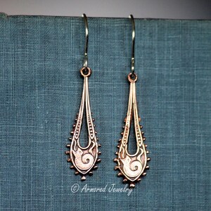 May include: A pair of copper-colored teardrop earrings with intricate filigree designs. The earrings have a long, slender shape with a decorative pattern at the bottom and a hook for the ear. The earrings are displayed against a teal background.