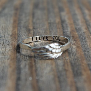 Puede incluir: Un anillo de plata con un dise&ntilde;o de un par de alas de &aacute;ngel. El anillo est&aacute; grabado con las palabras "Te amo".