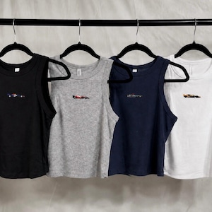 May include: Four sleeveless racerback tank tops in black, heather gray, navy blue, and white. Each tank top features a small, centered embroidered graphic of a Formula One race car on the chest. The garments are displayed on black hangers.