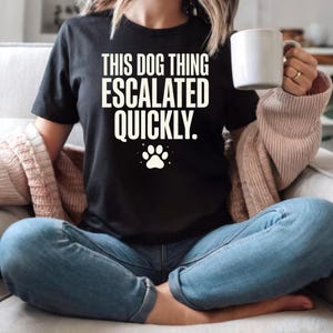 Puede incluir: Una camiseta negra de manga corta con el texto en blanco THIS DOG THING ESCALATED QUICKLY. y un gráfico de una huella de perro blanca centrado en el pecho. La prenda se combina con vaqueros azules y un cárdigan de punto beige.