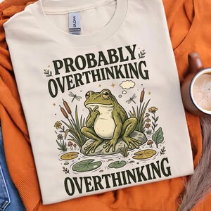 May include: A cream-colored t-shirt featuring a graphic of a green frog sitting on a rock in a pond. The frog has a hand to its chin in a thoughtful pose. The text "PROBABLY OVERTHINKING" appears above the frog, and "OVERTHINKING" is below.