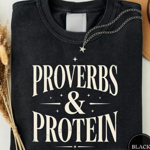 May include: A black crewneck sweatshirt featuring the white text "PROVERBS & PROTEIN" printed in a bold, serif font. The design includes small star accents above and below the text, with a thin silver chain necklace resting on the collar.