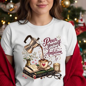 May include: A white t-shirt featuring a graphic of a silver moka pot pouring coffee into a cup. The design includes books, a pen, a candle, glasses, and flowers, with the text "Pour Yourself A Cup of Ambition" written in dark red script.