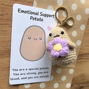 Könnte beinhalten: Ein gehäkelter Kartoffel-Schlüsselanhänger mit einer lila und gelben Blume, neben einer Karte mit der Aufschrift "Emotional Support Potato". Die Karte zeigt eine Cartoon-Kartoffel und den Text "You are a special potato. You are strong, you are loved, and you are enough."