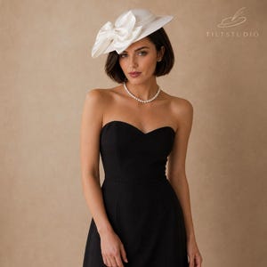May include: A white hat with a large bow, worn with a black strapless dress and a pearl necklace. The hat is the focal point, with a wide brim and a prominent bow on the side. The dress has a sweetheart neckline.