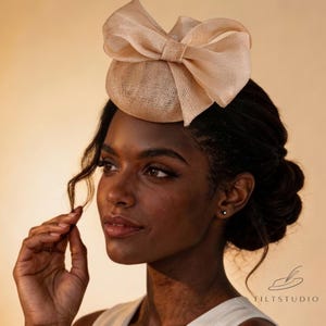 May include: A beige headpiece with a large bow. The headpiece is made of a textured fabric and sits atop a person's head. The background is a warm, neutral tone. The Tiltstudio logo is visible in the bottom right corner.