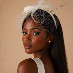 May include: Ivory-colored bridal headpiece with a bow and netting. The headpiece is worn on the head and has a delicate, vintage-inspired design. The netting covers the face, and the bow is positioned at the top of the head. The Tiltstudio logo is visible.