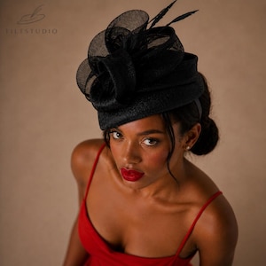 May include: A black formal hat with a layered design and feather accents. The hat is worn on the head and is made of a woven material. The hat is a stylish accessory for special occasions.