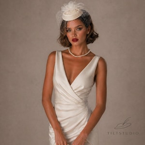 Pode incluir: Um chapéu creme, de estilo vintage, com véu, usado com um colar de pérolas e um vestido creme com decote em V. O chapéu tem uma base pequena e redonda e é adornado com um véu delicado e uma flor decorativa. O vestido tem um design cruzado.