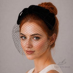Puede incluir: Una diadema de terciopelo negro con un lazo grande, combinada con un delicado velo de red negro. El velo está drapeado sobre la cara, creando un look de inspiración vintage. La diadema es un accesorio clásico.