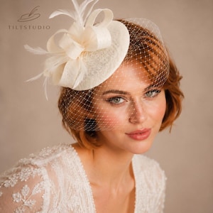May include: Cream-coloured fascinator featuring a bow and feathers, complemented by a fine veil. The headpiece is set against a neutral backdrop, with the Tiltstudio logo visible. A chic accessory for events.