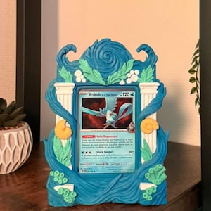 Peut inclure: Un cadre décoratif bleu avec une carte Pokémon à l'intérieur. Le cadre présente un motif de vagues avec des accents de coquillages. La carte présente une créature bleue en forme d'oiseau avec le texte "Artikodin" et "120".