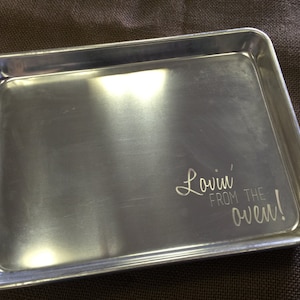 May include: A rectangular, silver-colored baking sheet with rounded corners. The phrase "Lovin' from the oven!" is etched on the lower right corner. The baking sheet is made of metal.
