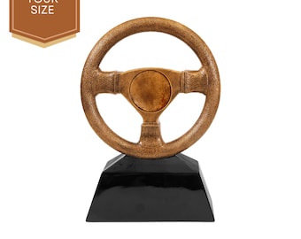 May include: A gold-coloured steering wheel trophy on a black base. The steering wheel has a textured surface and a round centre.