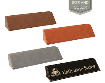 May include: A set of four desk nameplates in different colors. The nameplates are made of faux leather and have a triangular shape. The nameplate in the foreground is black with gold lettering and a logo that says "Mountain" and has a wave design. The nameplate is engraved with the name "Katharine Bates".