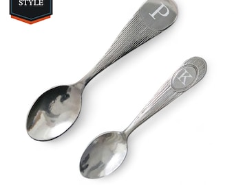 May include: Two silver spoons with engraved letters "P" and "K" on the handles. The spoons have a textured design and are laid out on a white background.
