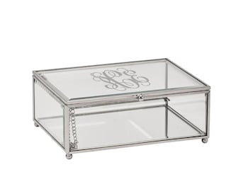 May include: A rectangular glass jewelry box with a silver metal frame and a hinged lid. The lid features an engraved monogram design. The box has small ball feet and a chain to hold the lid open.