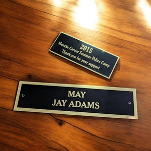 May include: Two gold plaques on a wooden surface. The top plaque is a dark gold and reads "2015 Moncks Corner Summer Police Camp Thank you for your support". The bottom plaque is a lighter gold and reads "MAY JAY ADAMS".