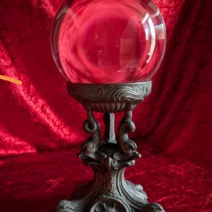Könnte beinhalten: Eine klare, kugelförmige Glaskugel auf einem dunklen, verzierten Metallständer. Der Ständer weist komplizierte, skulpturale Details mit geschwungenen, delfinartigen Figuren auf, die die Schale tragen, in der die glatte, transparente Glaskugel ruht.