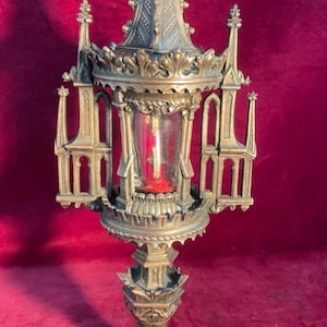 Puede incluir: Un relicario metálico con una compleja estructura arquitectónica en tonos bronce, con agujas puntiagudas y una cruz central en la parte superior. Un cilindro de cristal transparente en el centro revela un interior de tela roja y un objeto vertical.