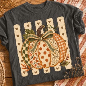 May include: A charcoal gray t-shirt featuring a central graphic of a pumpkin decorated with various patterns including polka dots, floral prints, and checkered sections. The pumpkin is tied with a sage green bow and sits against a background of vertical cream stripes adorned with small brown hearts.