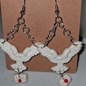 May include: White owl earrings with silver chain accents. Each earring features a detailed owl pendant with outstretched wings and a small envelope charm with a red seal. The earrings hang from silver fish hook ear wires.