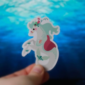 Pode incluir: Um adesivo de cavalo-marinho branco e verde com uma flor rosa e vermelha nas costas. O adesivo &eacute; segurado na m&atilde;o contra um fundo azul.