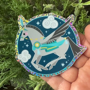 Puede incluir: Una pegatina brillante con un caballo alado blanco con una silla de montar azul y verde, volando sobre un fondo azul oscuro con nubes blancas y estrellas.