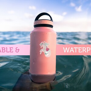 Pode incluir: Uma garrafa de &aacute;gua rosa com tampa e al&ccedil;a pretas. A garrafa tem um adesivo de sereia. A garrafa est&aacute; flutuando no oceano. O texto "DURABLE & WATERPROOF" est&aacute; em uma faixa rosa.