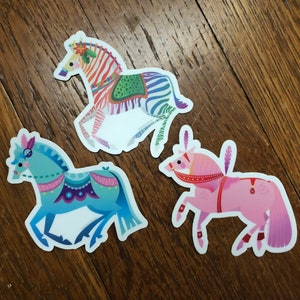 May include: Three colorful horse stickers with intricate details. The first horse is a zebra with green, purple, and white stripes. The second horse is blue with a white and purple saddle. The third horse is pink with a red and white saddle.