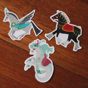 Pode incluir: Tr&ecirc;s adesivos coloridos com criaturas m&iacute;ticas: um cavalo alado com uma sela azul e verde, um cavalo preto com uma sela vermelha e dourada e um hipocampo com uma cauda rosa e verde.
