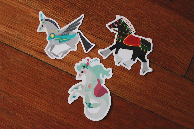 Pode incluir: Tr&ecirc;s adesivos coloridos com criaturas m&iacute;ticas: um cavalo alado com uma sela azul e verde, um cavalo preto com uma sela vermelha e dourada e um hipocampo com uma cauda rosa e verde.