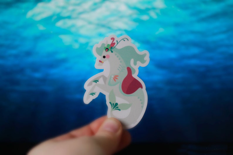 Pode incluir: Um adesivo de cavalo-marinho branco e verde com uma flor rosa e vermelha nas costas. O adesivo &eacute; segurado na m&atilde;o contra um fundo azul.