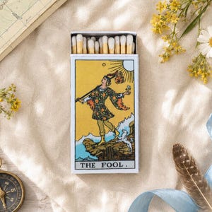 May include: A rectangular matchbox featuring a yellow illustration of The Fool tarot card on the front, with a figure carrying a bundle on a stick and a small white dog. The box is filled with wooden matches with white tips, arranged in a row.