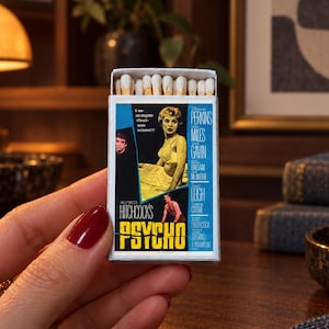 May include: A rectangular matchbox featuring a graphic print of the movie poster for Alfred Hitchcock's Psycho. The box displays the film title in yellow text, cast names, and scenes from the movie. A row of white-tipped wooden matches is visible.