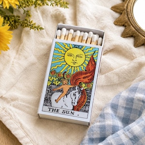 May include: A rectangular matchbox filled with wooden matches, featuring a colorful tarot card illustration of The Sun on the front. The design depicts a bright yellow sun with a face, a person riding a white horse, and sunflowers, with the text THE SUN.