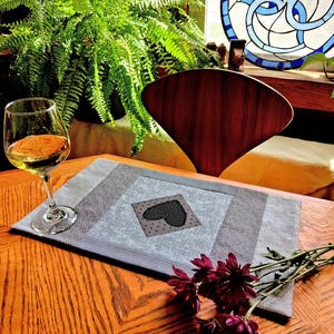 May include: A gray and light blue quilted placemat with a black heart design, a glass of white wine, and a bouquet of dark red flowers on a wooden table. A fern and a wooden chair are in the background.