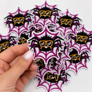 May include: A collection of embroidered patches featuring a black spider with orange-rimmed glasses centered on a white and magenta spiderweb. Each patch displays a round black spider body with eight legs and a small white mouth detail.