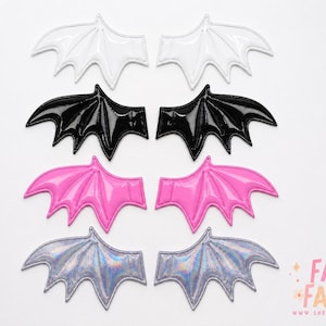 Può includere: Una collezione di toppe a forma di ali di pipistrello in vari colori, tra cui bianco, nero, rosa e argento iridescente. Le toppe sono disposte a coppie, mostrando il loro design simmetrico e la finitura lucida. L'immagine è su sfondo bianco.