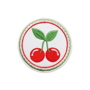 Puede incluir: Un parche bordado circular que presenta dos cerezas de color rojo brillante con tallos y hojas verdes sobre un fondo blanco. El diseño está encerrado por un borde circular rojo fino y un borde exterior decorativo con un patrón festoneado verde claro.