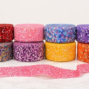 Könnte beinhalten: Acht Rollen dekorativen Bandes in verschiedenen Farben, darunter Rot, Rosa, Blau, Lila, Gelb und Orange. Das Band ist mit Pailletten bedeckt, und ein rosa Paillettenband ist im Vordergrund ausgerollt.
