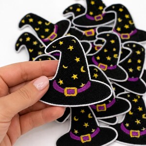 May include: A collection of embroidered witch hat patches featuring a black base with yellow stars and crescent moons. Each patch includes a purple hat band with a yellow buckle and a white stitched border, presented in a pile of multiple units.