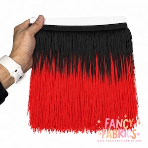 Puede incluir: Un fleco decorativo con una sección superior negra que transiciona a una sección inferior de color rojo vibrante. El fleco está unido a una banda de cabecera tejida negra. El texto FANCY FABRICS aparece en la esquina inferior derecha.