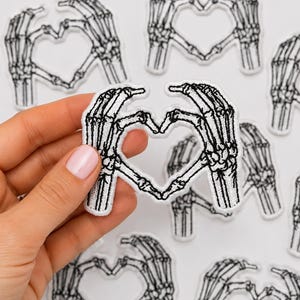May include: A white embroidered patch featuring a black skeleton hand design. The two hands are positioned to form a heart shape in the center. The patch has a clean white border and is shown against a background of multiple identical patches.