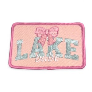Puede incluir: Un parche rectangular bordado con fondo rosa claro y borde rosa a juego. El parche presenta la palabra LAKE en letras mayúsculas de color azul claro, con un lazo rosa centrado encima y la palabra babe en letra cursiva debajo.